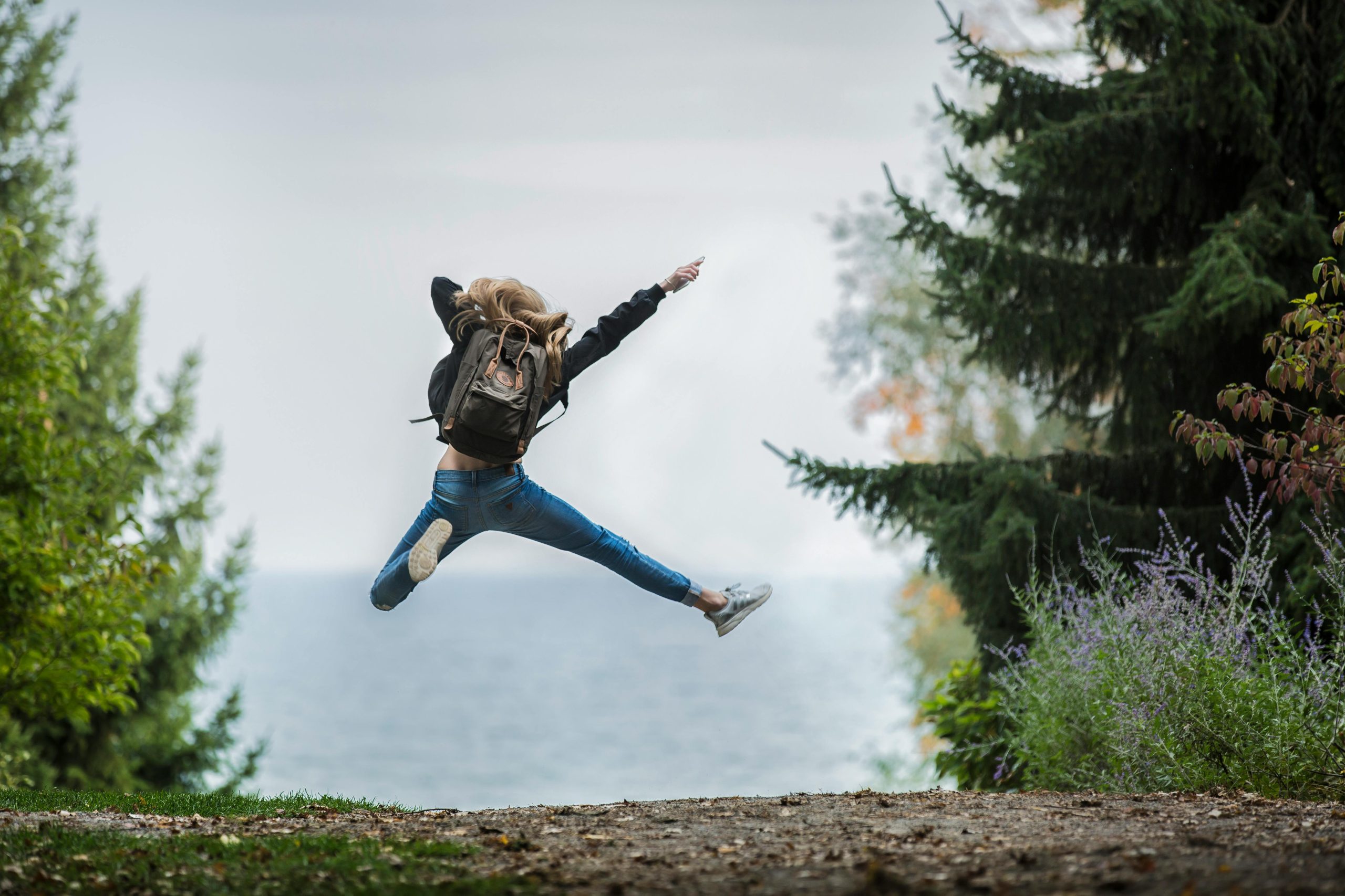 A long blonde haired woman jumping for joy. She is framed by trees on either side and a large hill in the background.